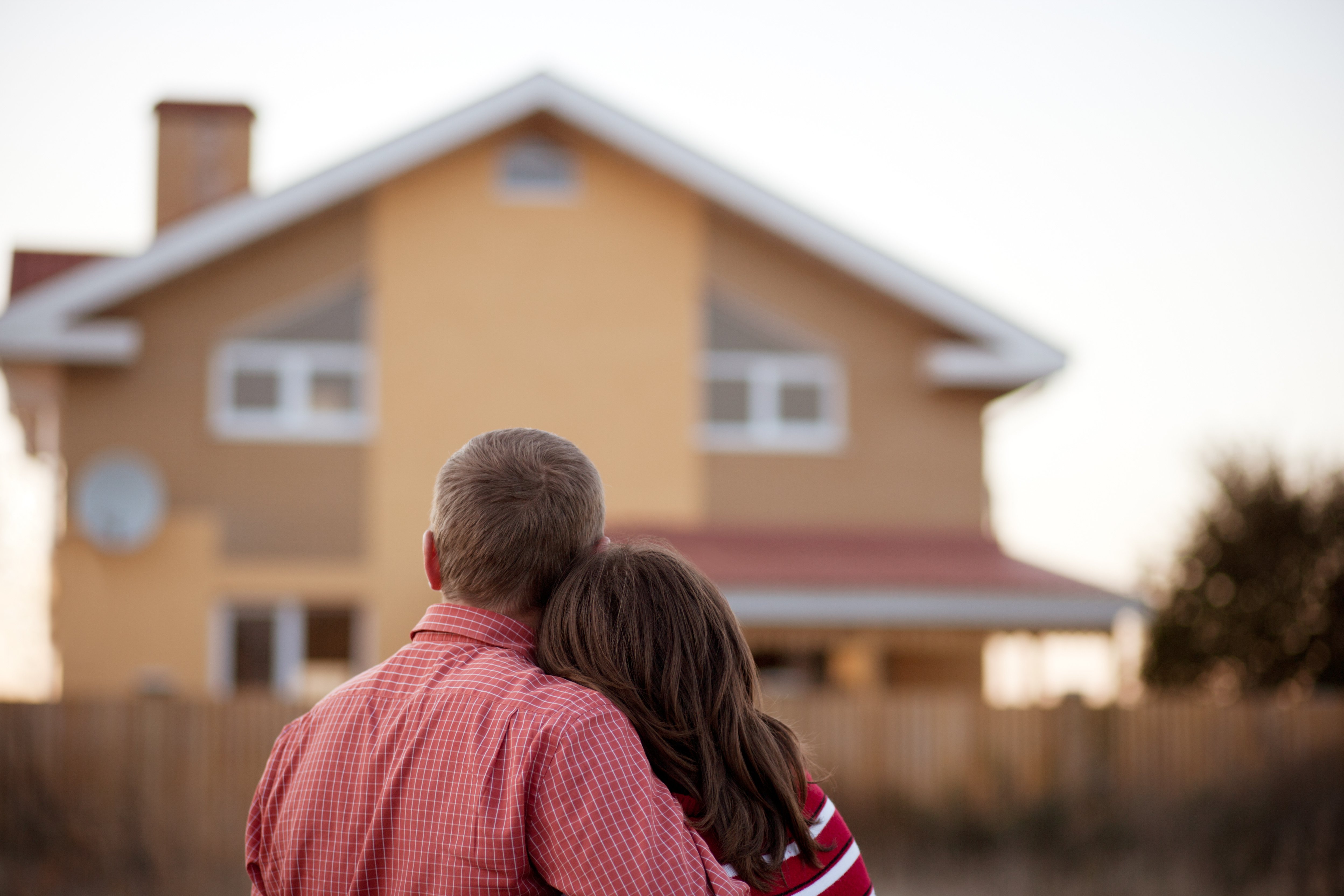  A couple stands together with their backs to the camera, looking at a house in the distance. The man has short blonde hair and is wearing a red plaid shirt, while the woman, with brown hair, is leaning her head on his shoulder, wearing a red and white striped shirt. The house in front of them is warm-toned, with a beige and light brown exterior, and appears to be in a peaceful, residential area. The image conveys a sense of togetherness, homeownership, and shared dreams.